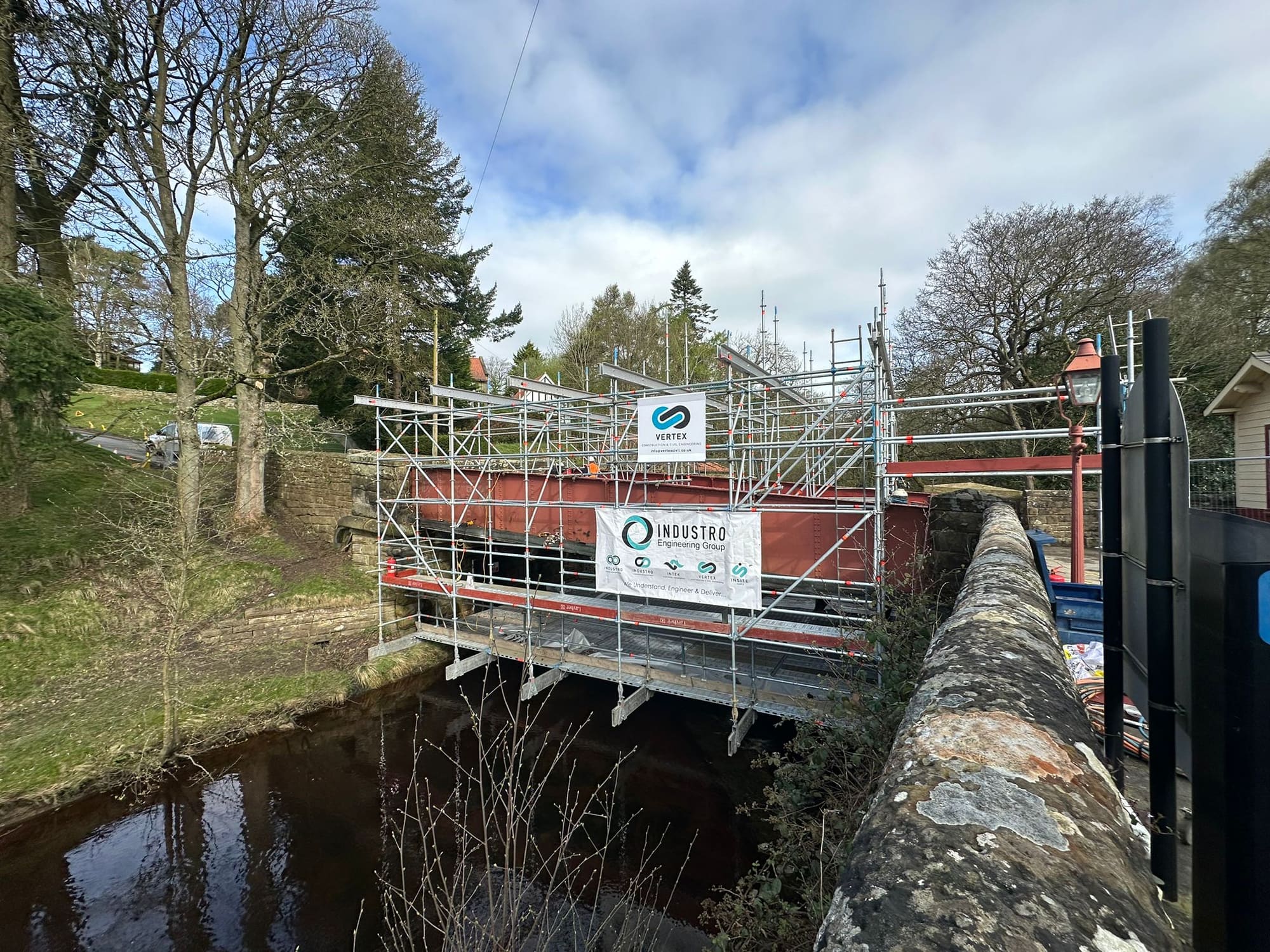 A railway bridge under refurbishment, fully scaffolded, with Vertex and INDUSTRO Engineering Group banners displayed